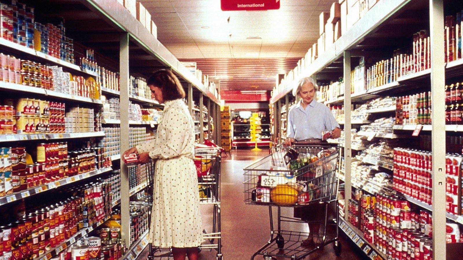 Two shoppers reading packages in a supermarket aisle lined floor to ceiling with canned and boxed brands.